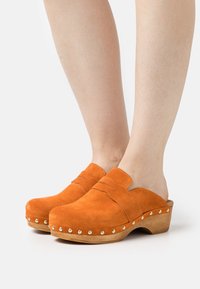 Personne portant des sabots en daim orange avec des semelles en bois et des clous dorés sur fond blanc.