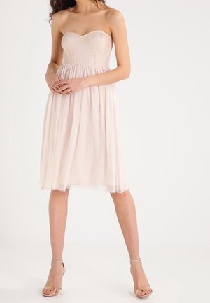 Béžové podkolenkové šaty bez ramínek, s krajkovým živůtkem a průsvitnou sukní, nošené s tělovými lodičkami na kotník, modelka stojí se sepjatýma rukama.