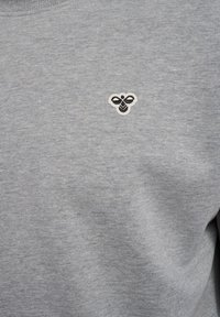 Gråt stof med en lille broderet sort-hvid bi-logo centreret på brystområdet af et stykke tøj.