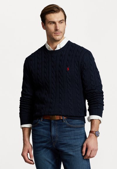 Marineblaue, kabelgewebte Strickjacke mit Rundhalsausschnitt und einem kleinen roten Logo. Kombiniert mit blauen Jeans und einem braunen Gürtel, dazu eine Uhr.