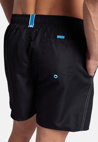 Bañadores negros con cinturilla elástica, raya de acento azul, bolsillo trasero con ojal azul y detalles de costura blancos.