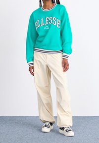 Sudadera turquesa con texto blanco "Ellesse", puños y dobladillo a rayas; combinada con pantalones de pierna ancha color crema y zapatillas negras.