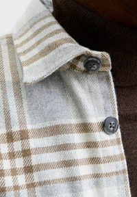 Camisa de franela a cuadros en tonos gris y beige con cuello abotonado, que presenta un suave cuello alto marrón debajo. Botones oscuros acentúan el diseño.