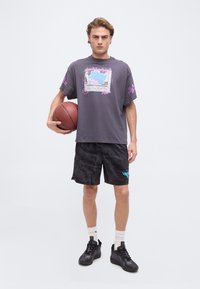 Νεαρός άνδρας που κρατάει ένα μπάσκετ, φοράει γκρι γραφικό t-shirt, μαύρα σορτς με σχέδια, λευκές κάλτσες και μαύρα αθλητικά παπούτσια, στέκεται σε απλο φόντο.