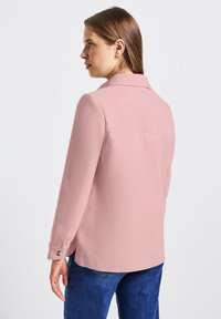 Světle růžová blůza s límečkem, dlouhými rukávy a knoflíkovými detaily na manžetách, v kombinaci s modrými denimovými džíny. Hladká textura.