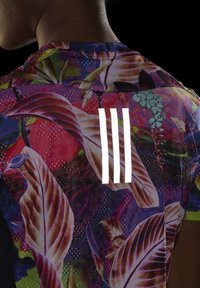 Osoba oblečená v blízkém záběru s sportovní košilí s multikolorovým květinovým vzorem a třemi vertikálními bílými pruhy na zádech, na tmavém pozadí.