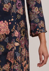 Blommönstrad klänning med en mörk marinblå bakgrund, med rosa, guld- och vita blommor. Materialet har en mjuk textur; trekvartslånga ärmar.