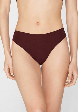 Vrouw draagt donkerbruine naadloze bikinibroekjes, staande met armen ontspannen langs haar zij tegen een effen achtergrond.