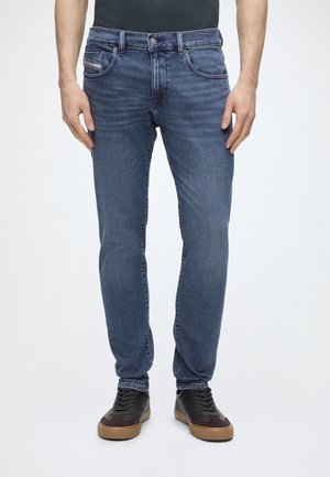 Homme portant un jean slim bleu et des baskets noires avec des semelles marron, debout devant un fond blanc uni.