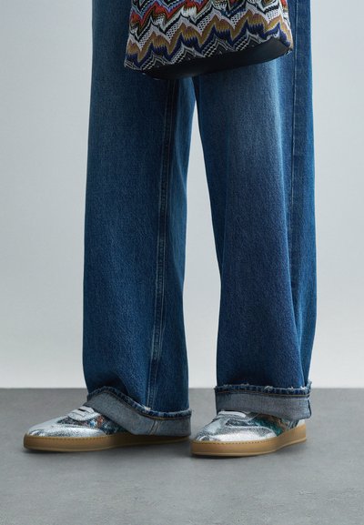 Des baskets argentées avec un design texturé, associées à un jean en denim bleu large. Les chaussures présentent une semelle lisse et des accents à motifs.