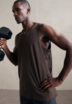 Débardeur de sport marron avec une coupe ample et un col rond. Un individu tient un haltère noir. Design simple, texture de tissu lisse, sans motifs.