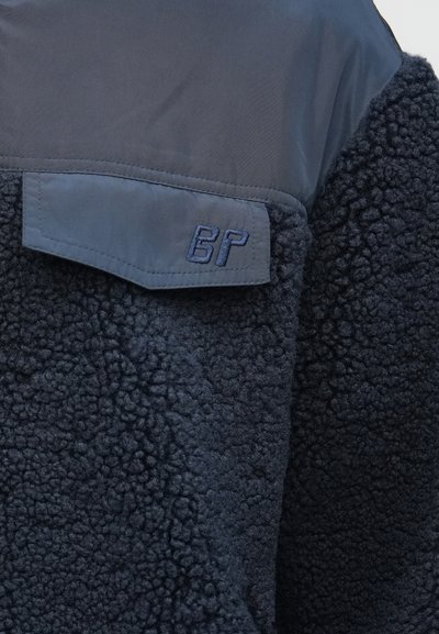 Σκούρο μπλε μπουφάν από φλις με υφή υφάσματος και μια λειανή τσέπη με καπάκι, κεντημένη με το "6P" στην περιοχή του άνω στήθους.