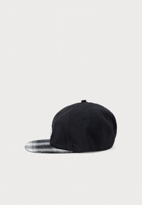 Casquette noire avec une visière plate à carreaux gris et blanc. Le matériau semble être en tissu avec un détail de logo sur le côté. Vue de côté affichée.