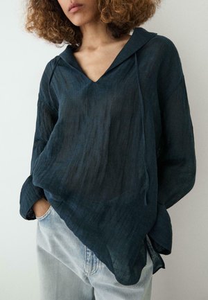 Kobieta ubrana w luźną, ciemnoniebieską bluzkę z kapturem o fakturowanej tkaninie oraz jasnoniebieskie dżinsy, pozująca z jedną ręką w kieszeni.