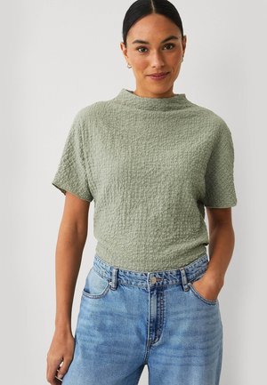 Vrouw draagt een lichtgroen textuur kort mouw shirt en een hooggetailleerde blauwe spijkerbroek, staand tegen een effen lichte achtergrond.