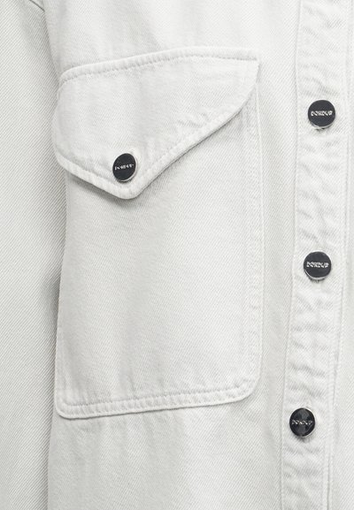 Κοντινό πλάνο από ανοιχτό γκρι ύφασμα denim με τσέπη με καπάκι, ασφαλισμένη με μεταλλικό κουμπί snap, και τρία μεταλλικά κουμπιά snap στη μπροστινή πλευρά του ενδύματος.