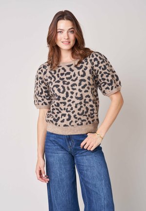 Vrouw met bruin haar die een beige en zwarte trui met korte pofmouwen en een luipaardprint draagt, en een blauwe spijkerbroek, staand met één hand in de zak.