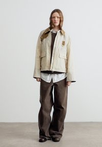 Νεαρό άτομο με μακριά μαλλιά στέκεται φορώντας ένα μπεζ σακάκι με καφέ γιακά, επάνω από λευκό πουκάμισο και σκούρο καφέ φαρδιά παντελόνια και παπούτσια.