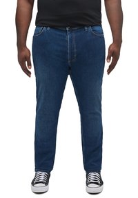 Jeans din denim albastru, cu croi drept, cinci buzunare, cusături vizibile și sistem de închidere cu nasture, asortați cu adidași negri și o cămașă neagră.