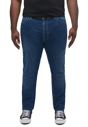 Vaqueros de mezclilla azul con corte recto, cinco bolsillos, costuras visibles y cierre de botón, combinados con zapatillas negras y una camisa negra.