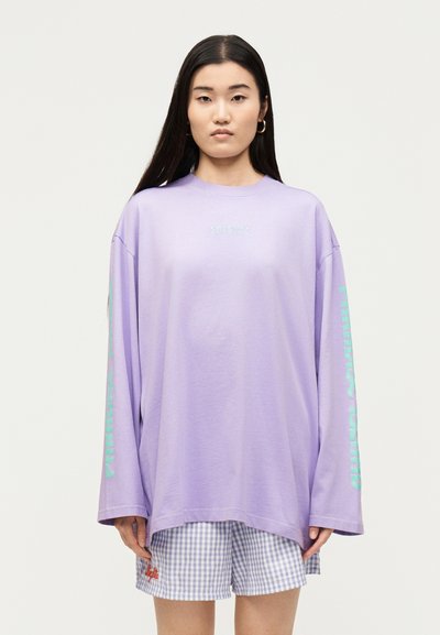 Γυναίκα με μακριά σκούρα μαλλιά που φοράει μια φαρδιά λεβαντίν μπλούζα με μακριά μανίκια με κείμενο στα μανίκια και καρό σορτς με πορτοκαλί λεπτομέρεια.