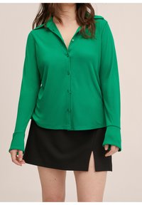 Mujer vestida con una blusa de botones de manga larga de color verde brillante y una falda mini negra con una apertura frontal, contra un fondo neutral.