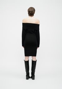 Kvinne med kort hår iført en off-shoulder svart kjole og knelengde svarte støvler, stående mot en ensfarget lys bakgrunn.