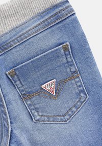 Jeans de mezclilla con una cinturilla acanalada gris, un parche con el logo en forma de triángulo en el bolsillo trasero y costuras visibles en un color contrastante.