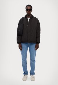 Sort, polstret jakke med knapper foran og elastiske manchetter, parret med lyseblå jeans og beige sneakers. Omfatter en sort skuldertaske og solbriller.