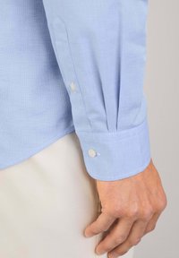 Camisa de vestir azul claro con un tejido texturizado, mangas largas y botones blancos en los puños. Ajuste entallado con dobladillo curvado.