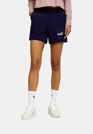 Persona che indossa pantaloncini Puma blu navy, felpa corta rosa chiaro, calzini bianchi Puma e sneakers beige con zeppa, in piedi con le mani nelle tasche.