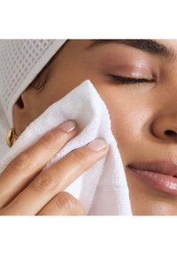 Primer plano de una mano sosteniendo una toalla blanca con textura contra la piel, con uñas naturales que presentan una sutil punta azul-verde. Tono de piel suave visible.