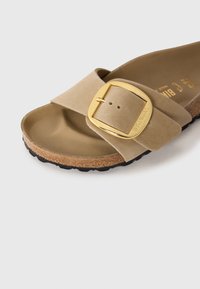 Bež suède Birkenstock sandala s kliznim krojem z velikim zlatim zaponko, korkovim podplatom in črnimi teksturiranimi podplati na beli podlagi.