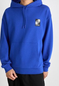 Blå hoodie med en framficka, justerbar dragsko och en svart och vit logopatch med ordet "Hugo". Slät tygkvalitet.
