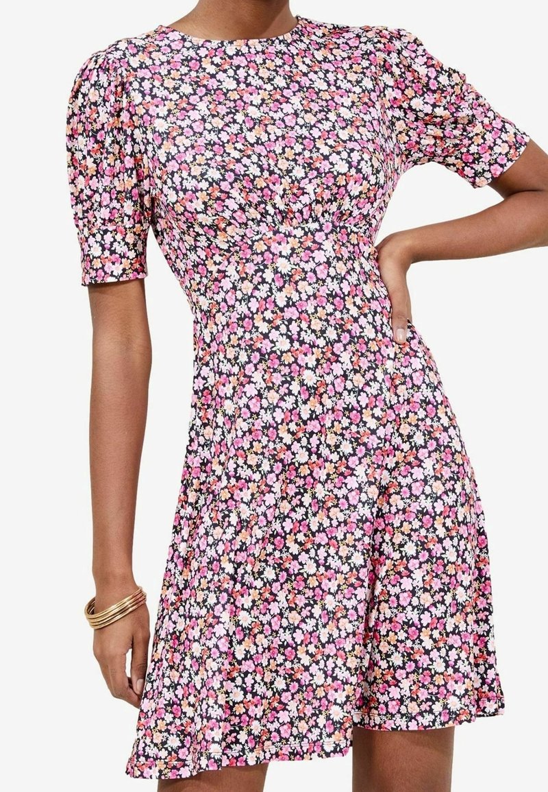 Femme portant une robe courte à fleurs roses, blanches et orange sur fond noir, avec des manches courtes bouffantes et des bracelets dorés au poignet gauche.
