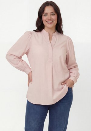 Smiling woman with dark hair wearing a light pink long-sleeve blouse and blue jeans, posing with one hand on hip and the other in pocket.
