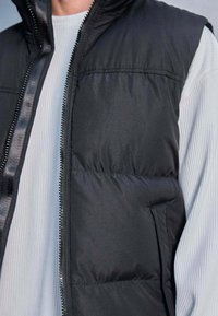 Gilet noir rembourré avec fermeture éclair à l'avant, poches latérales et texture lisse, superposé sur une chemise à manches longues légère à côtes.