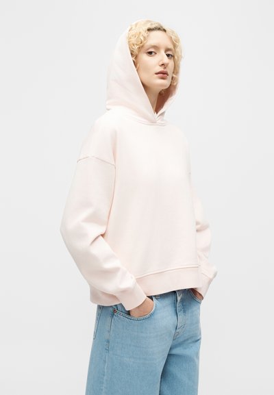 Νεαρό άτομο με σγουρά ξανθά μαλλιά που φοράει ροζ ανοιχτόχρωμο φούτερ με κουκούλα και ανοιχτό μπλε τζιν, στέκεται με τα χέρια στις τσέπες μπροστά σε ένα απλό φόντο.