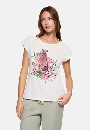 Mujer con una camiseta blanca que tiene un loro rosa con gafas de sol rodeado de flores, combinada con pantalones verde claro.
