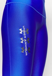 Siniset urheilulleggingsit kiiltävällä pinnalla. Niissä on hopeinen "Under Armour" -logo ja neljä mustaa yksityiskohtaa vasemmassa reidessä.