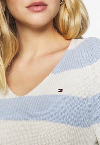 Maglione a maniche lunghe in maglia con righe orizzontali blu e crema, design a v, e un piccolo logo accentuato sul petto.