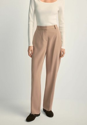 Kobieta ubrana w beżowe proste spodnie z wysokim stanem, biały prążkowany top z długim rękawem oraz ciemnobrązowe mokasyny, stojąca na neutralnym tle.