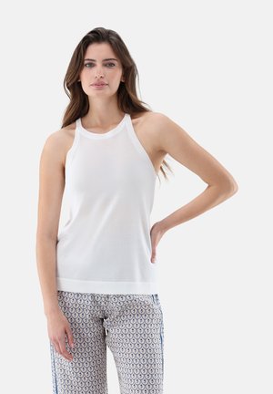 Frau mit langen braunen Haaren, die ein weißes ärmelloses Oberteil und gemusterte Hose trägt, steht mit einer Hand in der Hüfte vor einfarbigem Hintergrund.