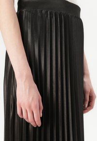 Jupe plissée noire en satin, dotée d'une ceinture lisse et d'une série de plis verticaux, mettant en valeur sa texture fluide.