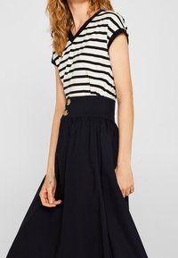 Top de manga corta a rayas en blanco y negro con escote en V, combinado con una falda negra de cintura alta y larga que presenta dos botones dorados.