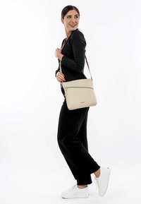 Sac bandoulière beige clair en matériau texturé, avec une fermeture éclair sur le dessus et une poche extérieure, associé à une tenue noire et des baskets blanches.