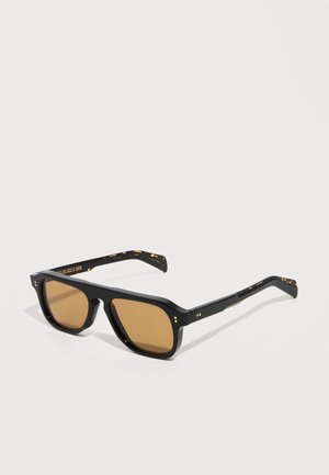 Mustad ristkülikukujulised päikeseprillid pruunikate läätsedega ning peenete kuldsete täppide aktsentidega raamidel ja külgede juures valgel taustal.