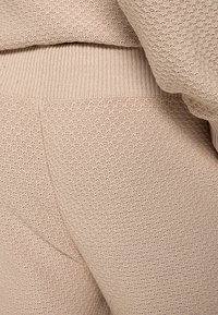 Beige, textured knit fabric featuring a loose weave, with ribbed accents at the waistband. Close-up of pants showcasing the material's pattern.
