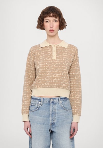 Beige zkrácená mikina s krémovým límcem, zdobená opakovaným ručně psaným vzorem. Měkká pletená textura, v kombinaci se světle modrými džíny s vysokým pasem.