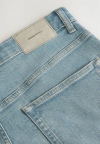 Jeans din denim de un albastru deschis, cu o etichetă rectangulară din piele gri inscripționată cu "DENIM SUPPLY" și detalii de cusătură vizibile.